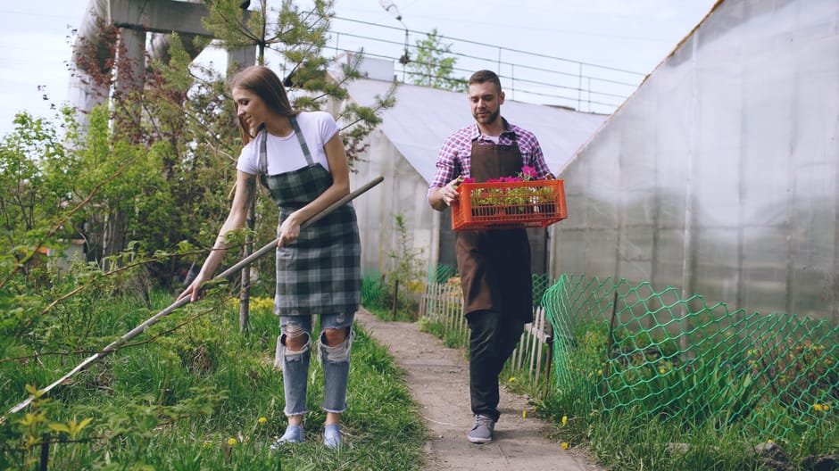 A young woman and a man are working together in a lush, green garden during daylight, with a clear sky overhead. The woman, dressed in a white t-shirt and ripped jeans, is using a long-handled garden 