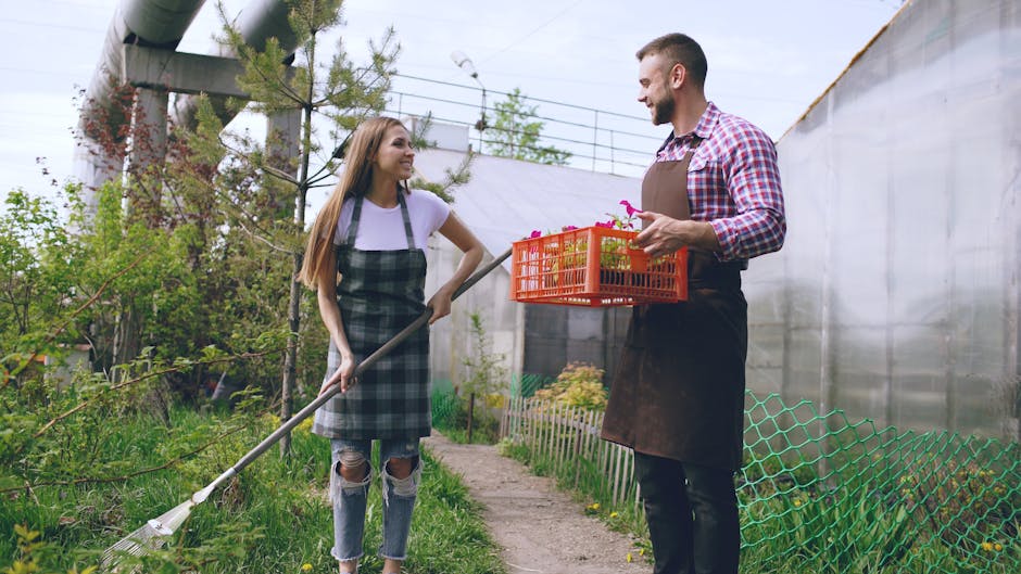 A young woman and a man are working together in a lush, green garden during daylight, with a clear sky overhead. The woman, dressed in a white t-shirt and ripped jeans, is using a long-handled garden 