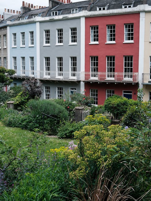 A multi-story residential building with a pastel blue and pink facade featuring numerous white-framed sash windows and small balconies with white railings. In the foreground, there is a lush, well-mai